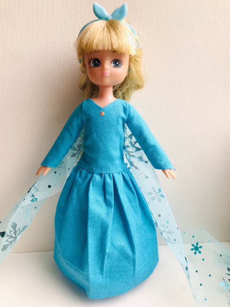 K&ouml;nnte beinhalten: Eine blaue und wei&szlig;e Puppe, die ein blaues Kleid mit Schneeflockenmuster und eine blaue Schleife im Haar tr&auml;gt.