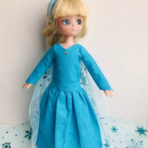 K&ouml;nnte beinhalten: Eine blaue Puppe mit blonden Haaren, die ein blaues Kleid mit einem T&uuml;llcape und einem blauen Schleifenhaarband tr&auml;gt. Die Puppe steht auf einem blauen und wei&szlig;en Hintergrund mit Schneeflockenmustern.