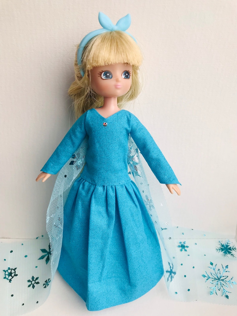 K&ouml;nnte beinhalten: Eine blaue Puppe mit blonden Haaren, die ein blaues Kleid mit einem T&uuml;llcape und einem blauen Schleifenhaarband tr&auml;gt. Die Puppe steht auf einem blauen und wei&szlig;en Hintergrund mit Schneeflockenmustern.