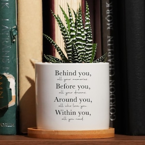 May include: A small potted succulent with green and white striped leaves sits on a wooden coaster. The white ceramic pot has text that reads "Behind you, all your memories..." The pot is on a shelf with a collection of books.