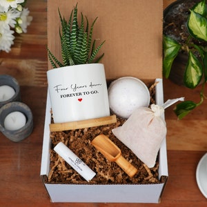 May include: A white gift box with a succulent plant in a white pot that says "Four Years Down FOREVER TO GO." The box also contains a bath bomb, a bag of bath salts, a wooden scoop, and a lip balm.