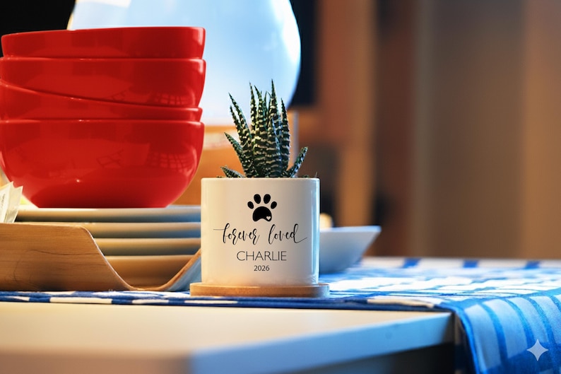 May include: A white cylindrical ceramic planter containing a small green succulent plant. The planter features a black paw print graphic above the text "forever loved", "CHARLIE", and "2026" printed in black ink on the front surface.