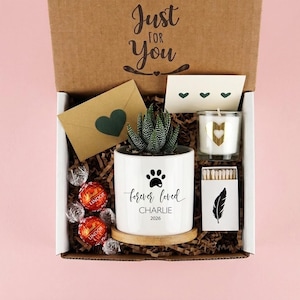 May include: A gift box containing a white ceramic planter with a succulent, a paw print, and the text 'forever loved CHARLIE 2026'. Includes a candle with a gold heart, a box of matches with a feather print, a brown envelope with a heart, and chocolates.