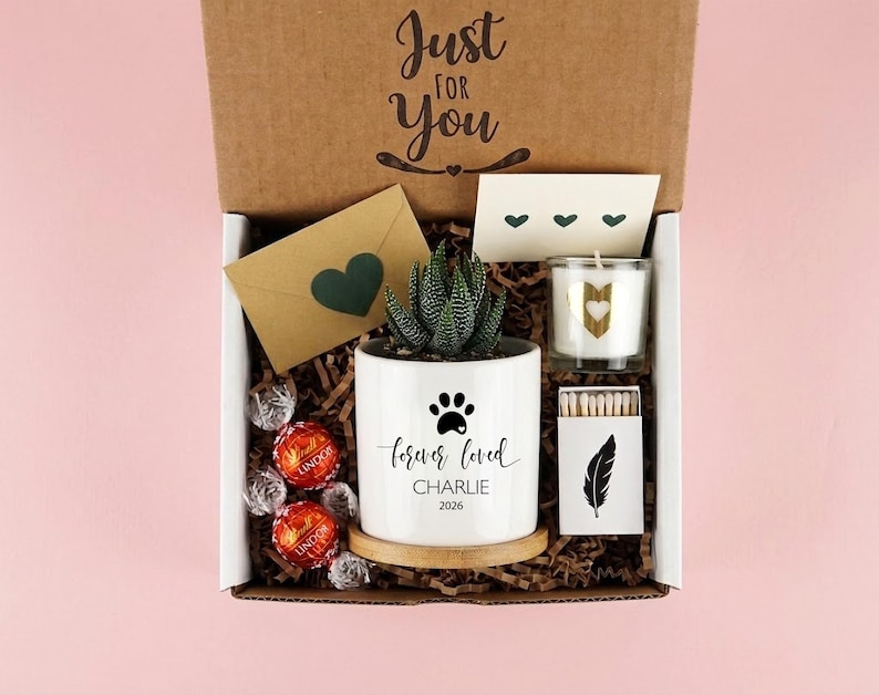 May include: A gift box containing a white ceramic planter with a succulent, a paw print, and the text 'forever loved CHARLIE 2026'. Includes a candle with a gold heart, a box of matches with a feather print, a brown envelope with a heart, and chocolates.