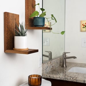 Op de afbeelding: Twee L-vormige houten wandplanken met een ingegraveerd bladpatroon op het oppervlak. De onderste plank bevat een witte pot met een gestreepte vetplant, de bovenste plank een blauwgroene pot met een hangende plant.