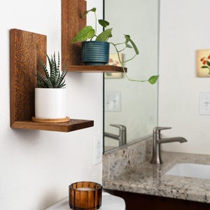 Op de afbeelding: Twee L-vormige houten wandplanken met een ingegraveerd bladpatroon op het oppervlak. Op de ene plank staat een witte pot met een gestreepte vetplant, op de andere een geribbelde blauwgroene pot met een hangende plant.