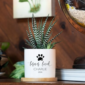 May include: A white cylindrical ceramic planter containing a green and white striped succulent plant. The planter features a black paw print graphic above the text "forever loved", the name "CHARLIE", and the year "2026" in black lettering.