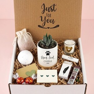 May include: A gift box containing a succulent in a white pot with a paw print and the text 'forever loved CHARLIE 2026', a bath bomb, a candle, a small pouch, a lip balm, a box of matches with a feather, a coffee packet, and two chocolates.