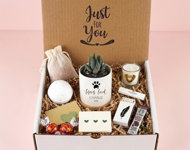 May include: A gift box containing a succulent in a white pot with a paw print and the text 'forever loved CHARLIE 2026', a bath bomb, a candle, a small pouch, a lip balm, a box of matches with a feather, a coffee packet, and two chocolates.