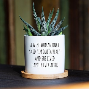 May include: A white cylindrical ceramic planter containing a green succulent plant with white speckled leaves. The planter features black printed text that reads: A WISE WOMAN ONCE SAID “IM OUTTA HERE” AND SHE LIVED HAPPILY EVER AFTER. It sits on a round bamboo tray.