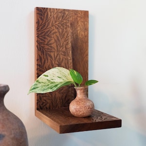 May include: A wall-mounted L-shaped wooden shelf featuring a dark brown finish with an engraved leaf pattern. A small, round, textured tan ceramic vase sits on the shelf, holding a single variegated green and white pothos plant leaf.