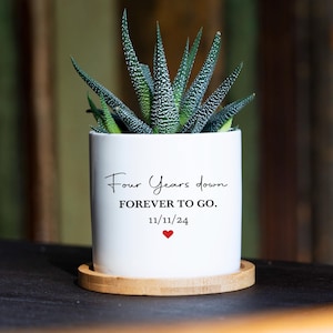 May include: A white cylindrical ceramic planter containing a green succulent plant. The planter features the black text "Four Years down FOREVER TO GO." and the date "11/11/24" above a small red heart. It sits on a circular light wood base.