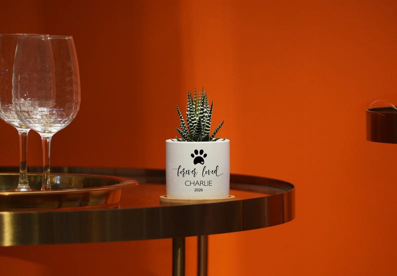 May include: A white cylindrical ceramic planter containing a small green and white striped succulent plant. The planter features a black paw print icon above the text "forever loved" followed by the name "CHARLIE" and the year "2026" in black.