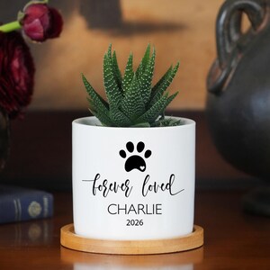 May include: A white cylindrical ceramic planter with a bamboo saucer containing a green succulent. The planter features a black paw print icon, the text "forever loved", the name "CHARLIE", and the year "2026" printed in black ink on the front.