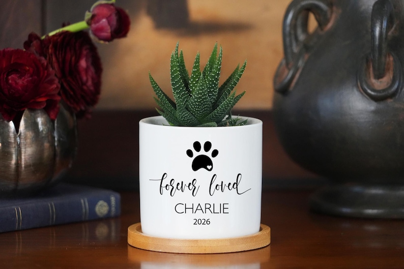 May include: A white cylindrical ceramic planter with a bamboo saucer containing a green succulent. The planter features a black paw print icon, the text "forever loved", the name "CHARLIE", and the year "2026" printed in black ink on the front.