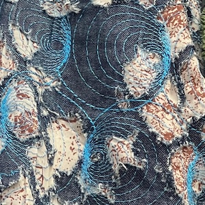 May include: A textile fabric featuring a layered design with dark blue denim patches and a cream-colored fabric with brown patterns underneath. The surface is decorated with bright blue circular machine embroidery stitching and frayed edges.