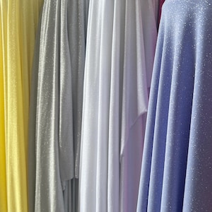 May include: A collection of draped fabric bolts in yellow, silver, white, and light purple. Each piece of material features a fine, shimmering glitter finish that catches the light, creating vertical folds and soft shadows across the surface.