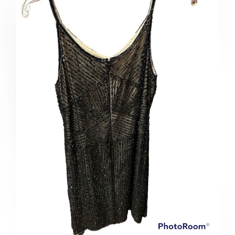 Puede incluir: Un vestido negro con cuentas y tirantes finos, colgado de una percha. El vestido presenta un patr&oacute;n geom&eacute;trico de cuentas y un escote en V. El vestido es sin mangas y tiene un cierre de cremallera en la espalda.