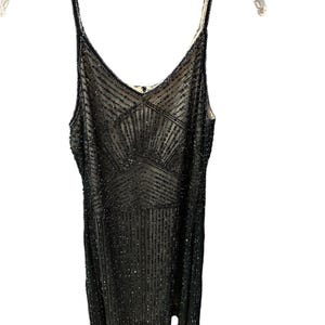 Puede incluir: Un vestido negro con cuentas con escote en V y tirantes finos. El vestido presenta un patr&oacute;n geom&eacute;trico en el corpi&ntilde;o y l&iacute;neas verticales de cuentas. El vestido est&aacute; colgado de una percha blanca.