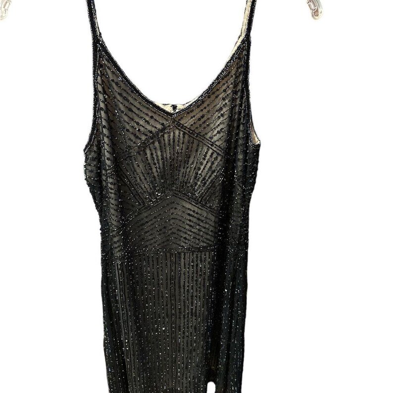 Puede incluir: Un vestido negro con cuentas con escote en V y tirantes finos. El vestido presenta un patr&oacute;n geom&eacute;trico en el corpi&ntilde;o y l&iacute;neas verticales de cuentas. El vestido est&aacute; colgado de una percha blanca.