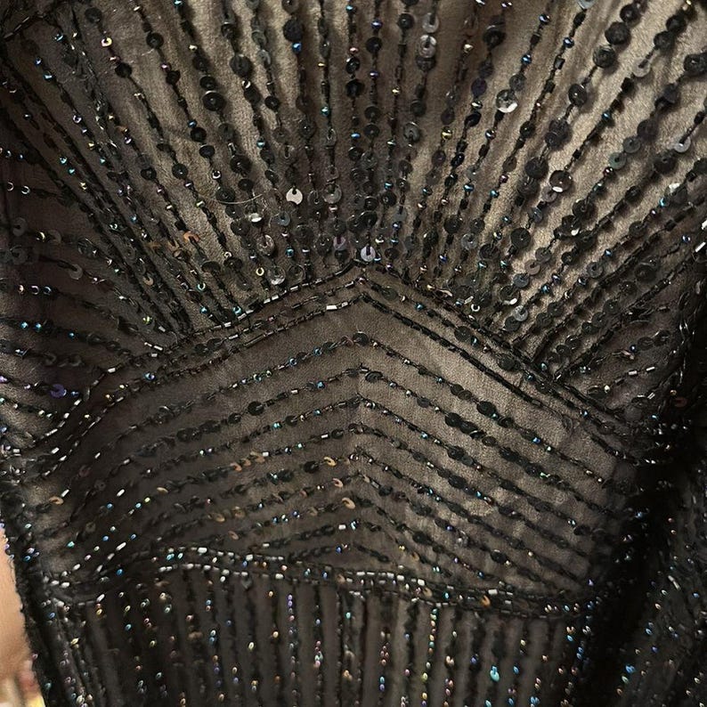 Puede incluir: Primer plano de un corpi&ntilde;o de vestido negro adornado con lentejuelas y cuentas. El dise&ntilde;o presenta patrones verticales y en zigzag, creando un efecto brillante. La tela parece ser un material oscuro y transparente.