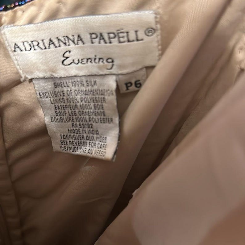 Puede incluir: Primer plano de una etiqueta de un vestido Adrianna Papell Evening. La etiqueta blanca presenta texto negro que detalla la composici&oacute;n de la tela del vestido, incluyendo seda y poli&eacute;ster. La etiqueta tambi&eacute;n indica que el vestido fue hecho en India.