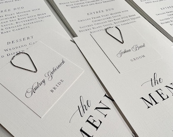 May include: A stack of white wedding menu cards with black text. The top card has a silver heart-shaped paper clip holding it to the card below. The text on the top card reads "the MENU" and includes the names "Audrey Gubernich" and "Joshua Beard" with the titles "Bride" and "Groom".