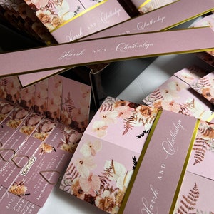 May include: A stack of folded wedding invitations with a floral design and gold accents. The invitations are pink with a gold ribbon and the names "Harsh and Chathushya" are printed in white cursive.