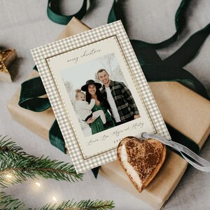 May include: A Christmas card with a family photo, featuring a gingham border and the words "merry christmas." The card is on a wrapped gift with a green ribbon and a gold heart ornament.