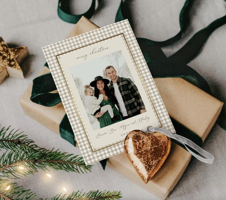 May include: A Christmas card with a family photo, featuring a gingham border and the words "merry christmas." The card is on a wrapped gift with a green ribbon and a gold heart ornament.