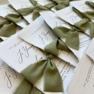 May include: A collection of white wedding invitation cards with deckled edges, each featuring black printed text and tied with a sage green silk ribbon. The text includes the names Ji Young and Leon, and the location Keswick Vineyards, Keswick, Virginia.