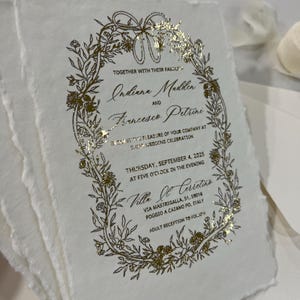 May include: Gold foil wedding invitation with a floral wreath design. The invitation reads "Together with their families, Indiana Madden and Francesco Petrini request the pleasure of your company at their wedding celebration. Thursday, September 4, 2025 at five o'clock in the evening. Villa Ol Cerretine, Via Mastrigalla, 51, 59016 Poggio a Cajano PO, Italy. Adult reception to follow."