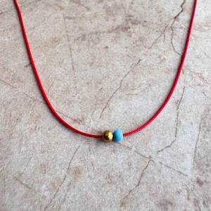 May include: A red string necklace with a gold and blue bead. The necklace is on a light brown surface.