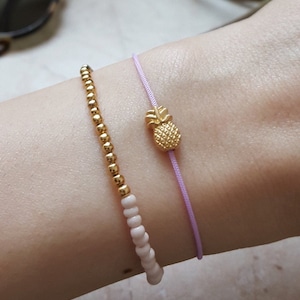 May include: Two bracelets on a wrist. One is a delicate gold chain with small beads. The other is a thin lavender cord with a gold pineapple charm.