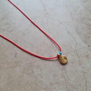 Può includere: Una collana di corda rossa con un ciondolo a spirale dorata e due piccole perle blu.