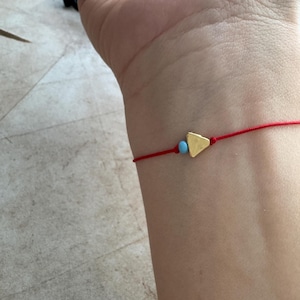 Könnte beinhalten: Rotes Armband mit einem goldenen Dreiecksanhänger und einer kleinen türkisfarbenen Perle. Das Armband ist schlicht und filigran und für das Handgelenk bestimmt.
