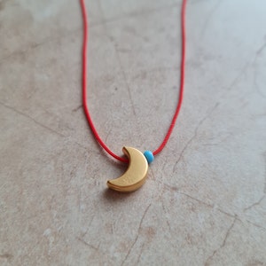 May include: A gold crescent moon charm hangs from a red string necklace with a small blue bead.