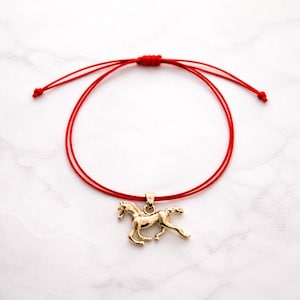 May include: A red string bracelet featuring a gold-coloured horse pendant. The horse is shown in a running pose. The bracelet is adjustable with sliding knots. The background is a white marble surface.