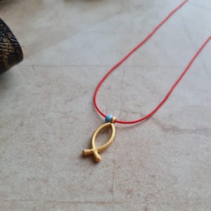 Op de afbeelding: Een gouden vis-hangerketting aan een rood koord met een lichtblauwe kraal.