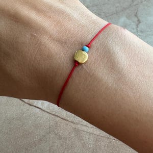 Può includere: Un braccialetto di corda rossa con un piccolo ciondolo dorato e una perla blu.