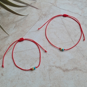 May include: Two red string bracelets with gold and turquoise beads. The bracelets are tied in a knot and are on a light brown surface.