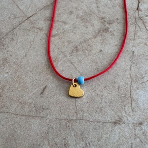 Op de afbeelding: Een rode koordketting met een klein gouden hartje en een blauwe kraal.