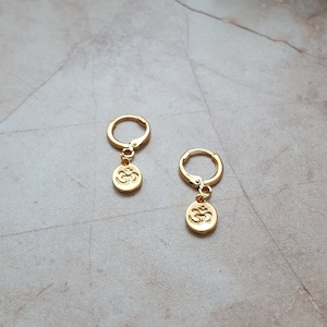 May include: Gold hoop earrings with a small Om symbol charm hanging from each hoop.