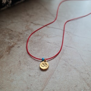 Op de afbeelding: Een gouden hangerketting met een rode koord. De hanger toont het Om-symbool en is geaccentueerd met een kleine blauwe kraal. De ketting wordt gepresenteerd op een neutraal getinte ondergrond.