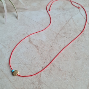May include: A red string necklace with a small gold charm. The charm has a blue stone in the center.