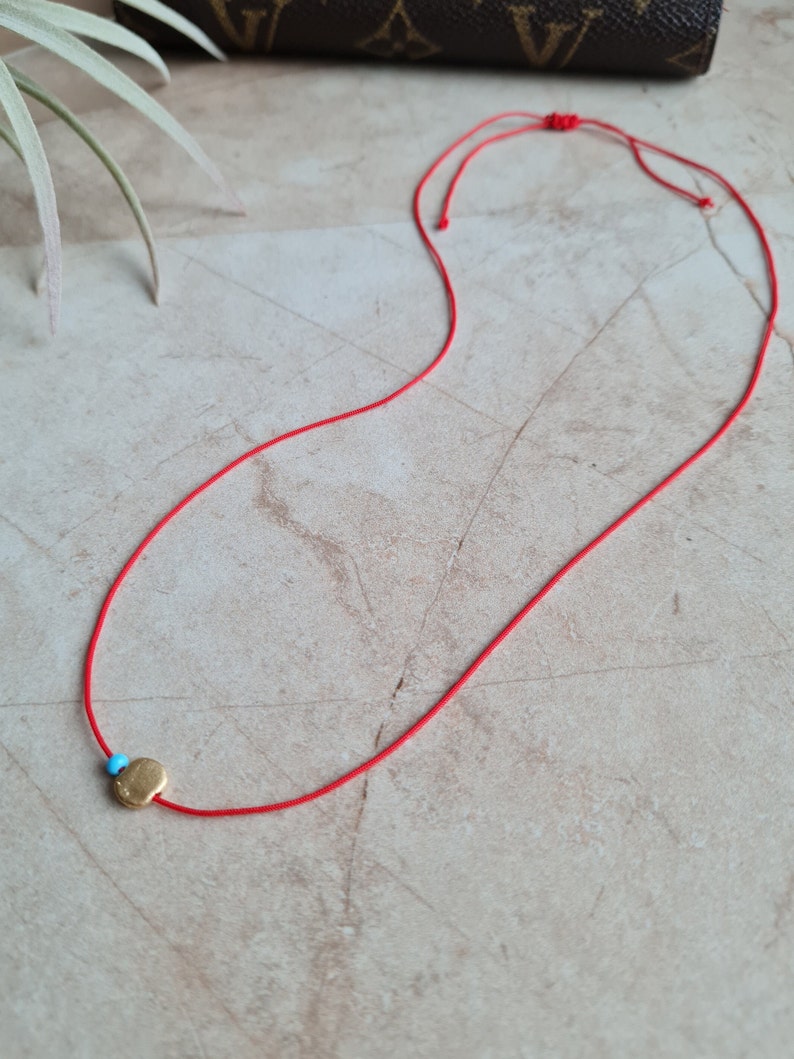 May include: A red string necklace with a small gold charm. The charm has a blue stone in the center.