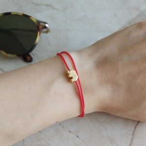 May include: A red string bracelet featuring a small gold-coloured elephant charm. The bracelet is worn on a wrist. Sunglasses are visible in the background. The elephant charm is central on the red string.