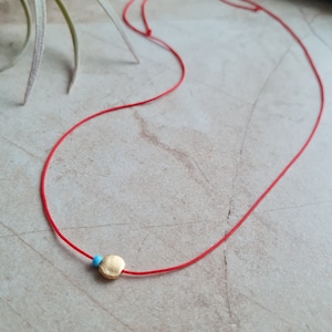 May include: A red string necklace with a small gold circle pendant and a blue bead.
