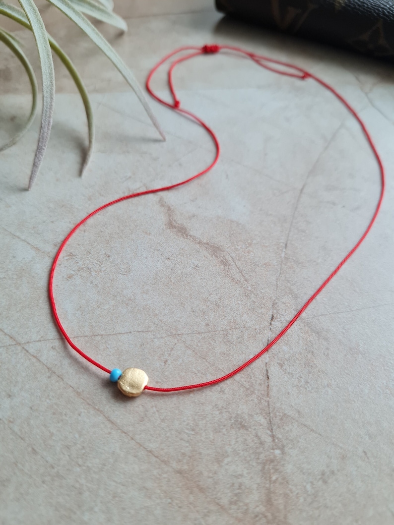 May include: A red string necklace with a small gold circle pendant and a blue bead.