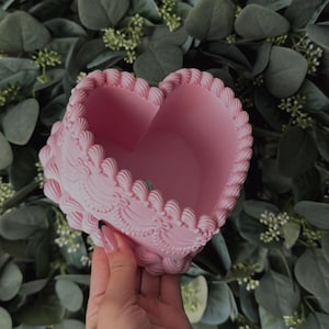 Pode incluir: Um vaso de coração rosa com um design decorativo e ornamentado. O vaso é feito de um material liso e brilhante.